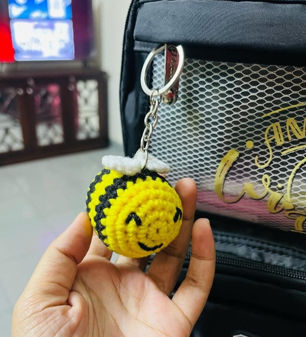 Yellow and black crocheted bumble bee bag charm with white wings and embroidered smiling face, attached to a backpack and held in a hand