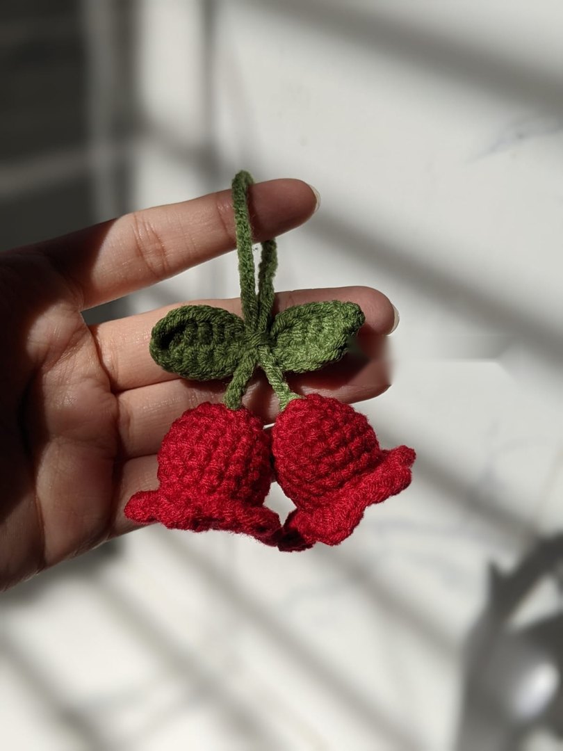 Pair of red crocheted lily of the valley flowers with green leaves and a green chain cord, held in a hand under bright window light