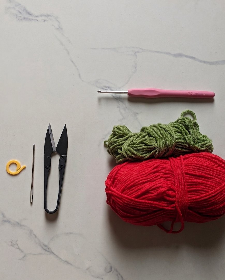 Materials flat lay for the lily of the valley crochet pattern: red and green milk cotton yarn, a 3 mm crochet hook, scissors, tapestry needle and stitch marker on white marble