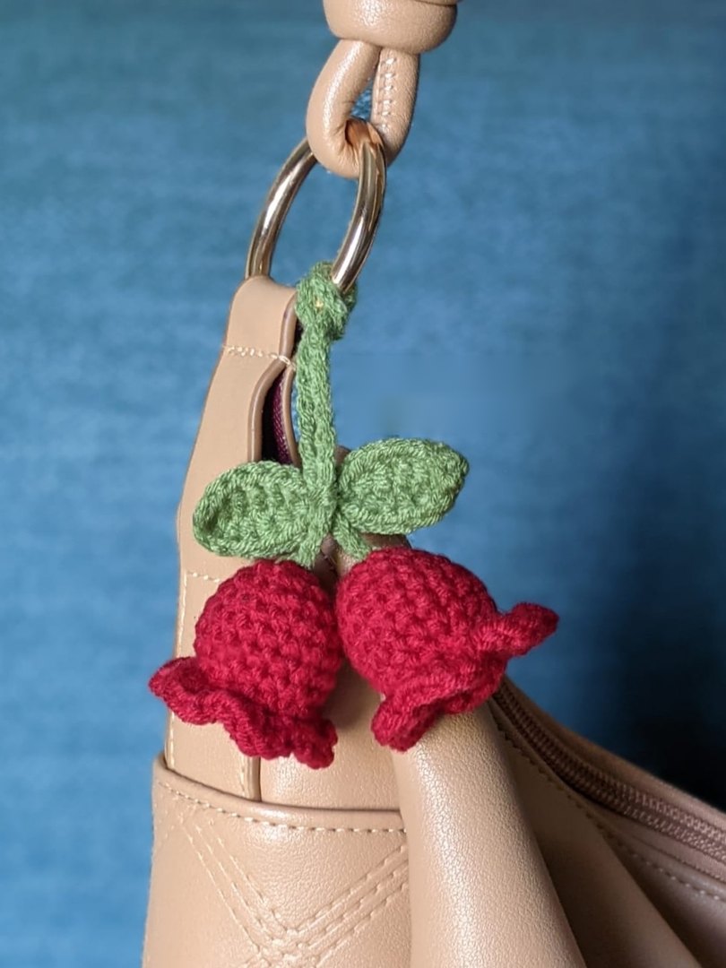 Two red lily of the valley crochet bag charms with a green chain cord and crocheted leaves, attached to a tan handbag with a metal split ring