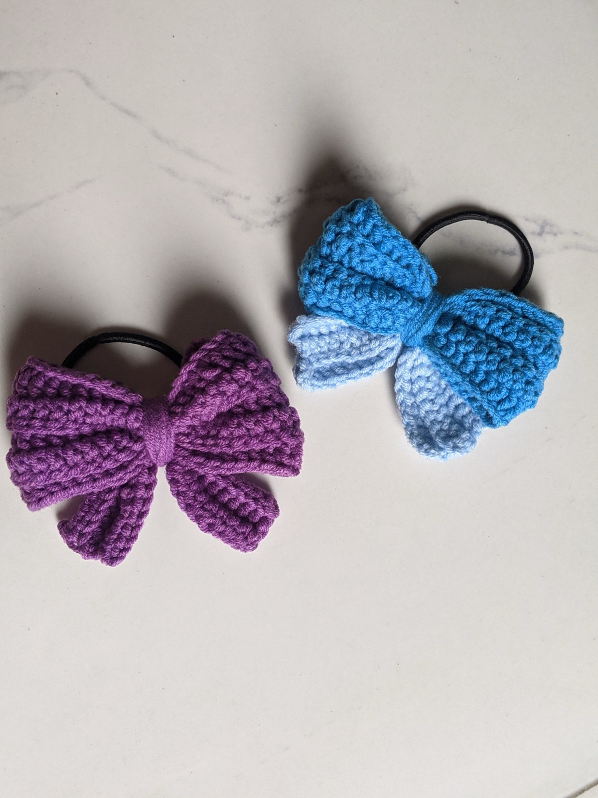Two finished crocheted puffy bow hair ties, purple on left and blue and light blue on right, photographed top-down on a white marble surface