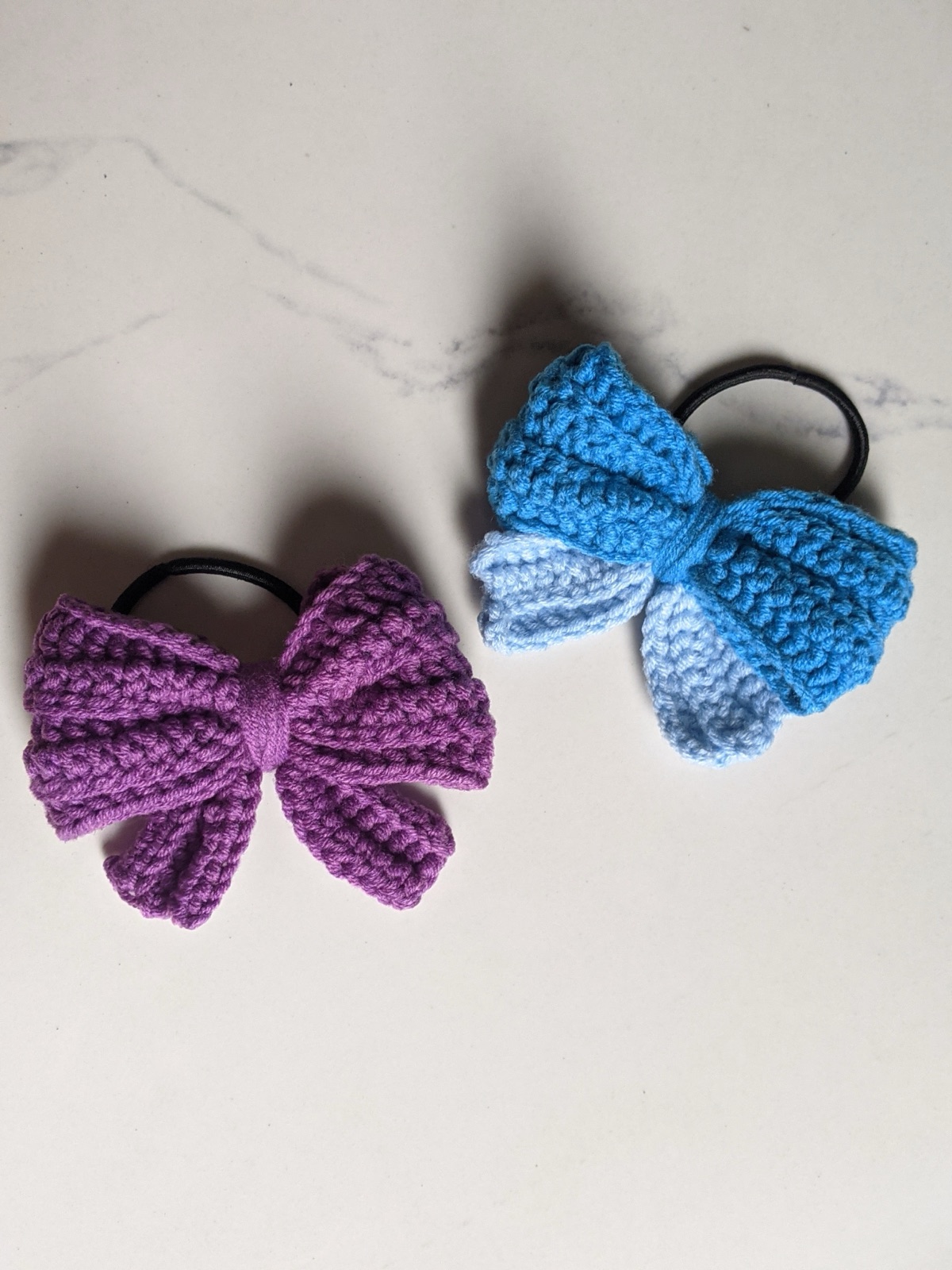 Two finished crocheted puffy bow hair ties, purple on left and blue on right, photographed on a clean white marble surface