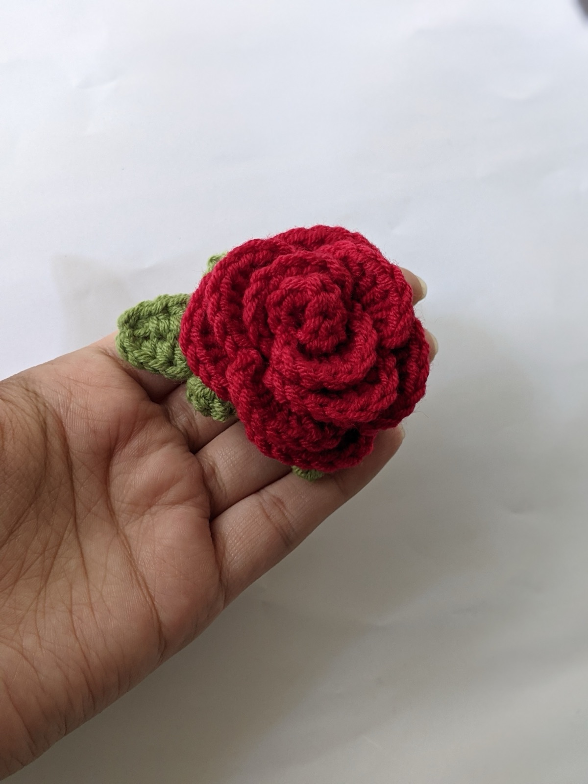 Crocheted red rose gajra hand flower with green leaves held in an open hand showing palm-size scale