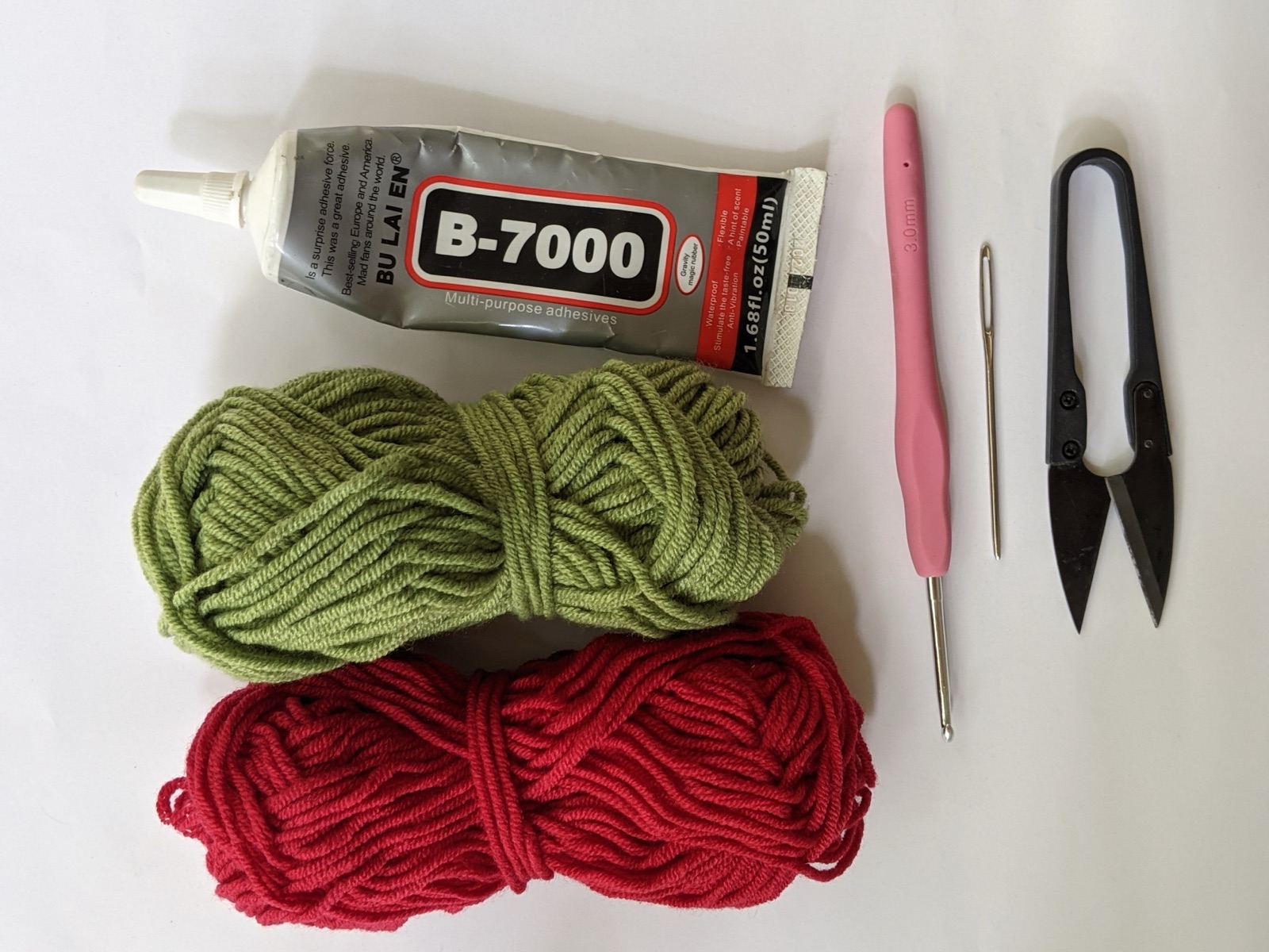 Materials flat lay for the rose gajra crochet pattern: red and green 4-ply milk cotton yarn, 3 mm pink crochet hook, tapestry needle, scissors, and B-7000 glue