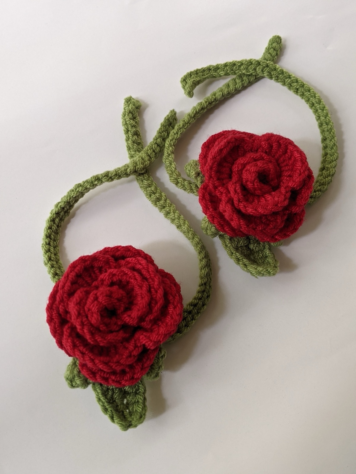 Pair of finished red rose crochet gajras with green leaves and tie strings laid out on a white marble background