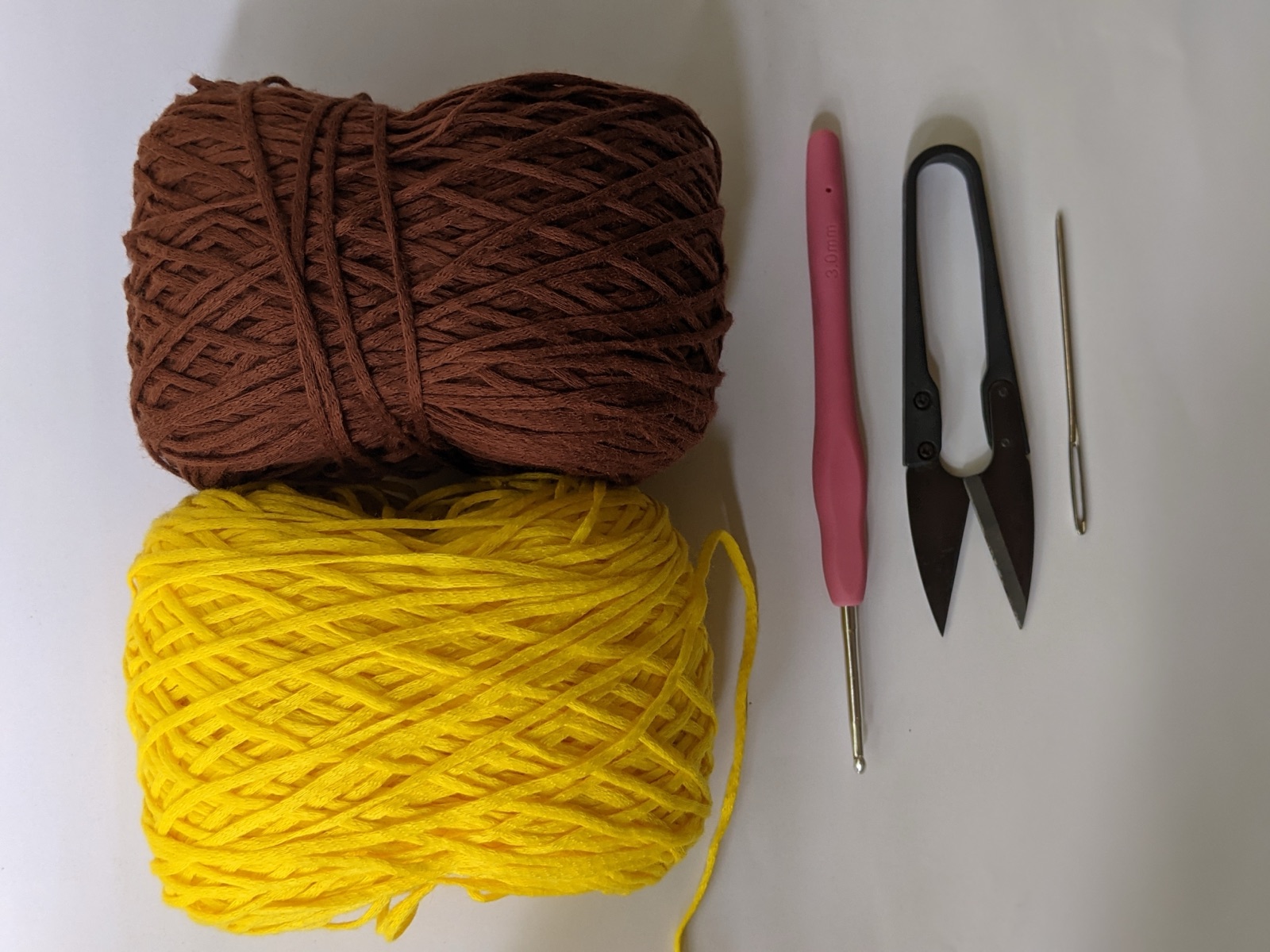 Materials flat lay for the sunflower coaster crochet pattern: balls of dark brown and yellow medium-weight yarn, a 3 mm pink crochet hook, scissors, and a tapestry needle