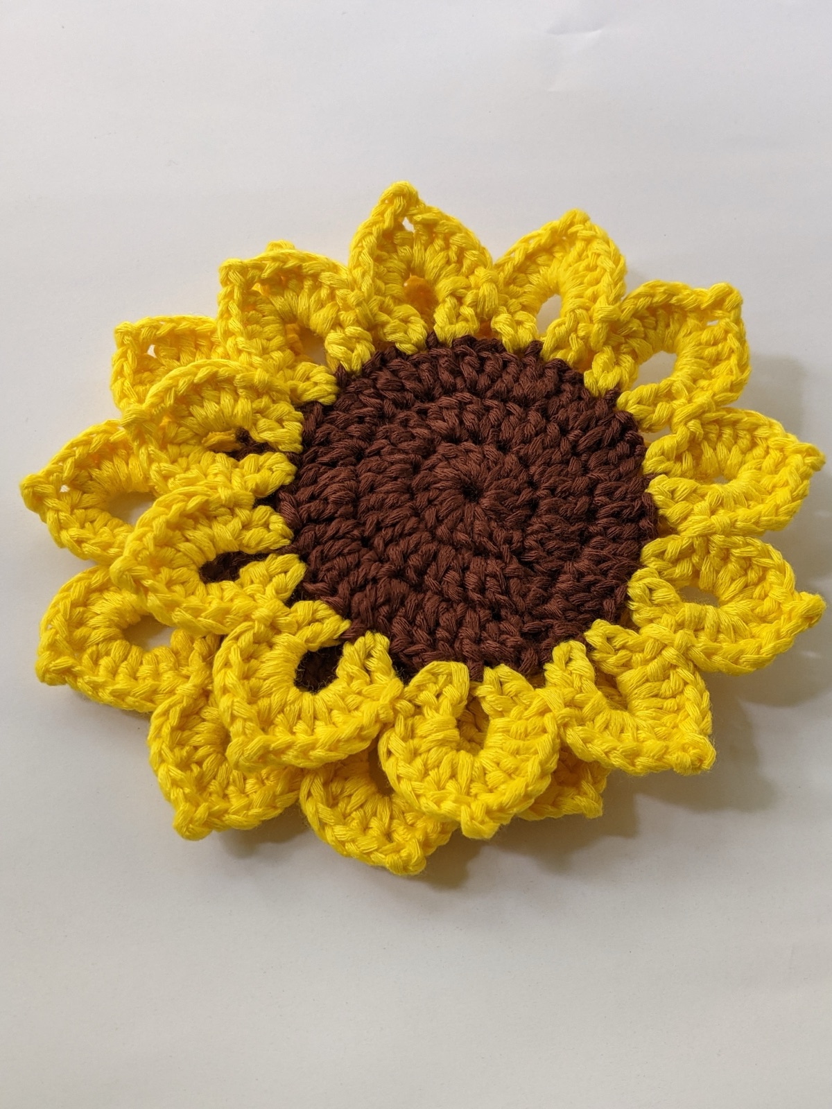 Single crocheted sunflower coaster tilted on a white surface with 11 fluffy yellow petals around a dark brown center