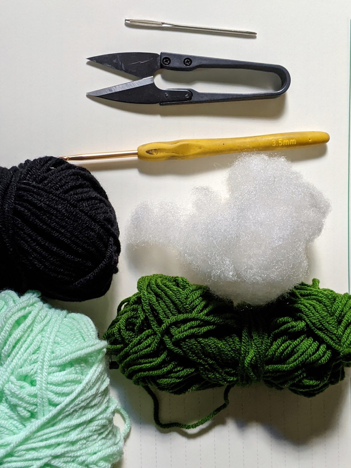 Flat lay of crochet supplies: dark green and mint green milk cotton yarn, black yarn, 3.5mm crochet hook, fiberfill stuffing, tapestry needle, scissors