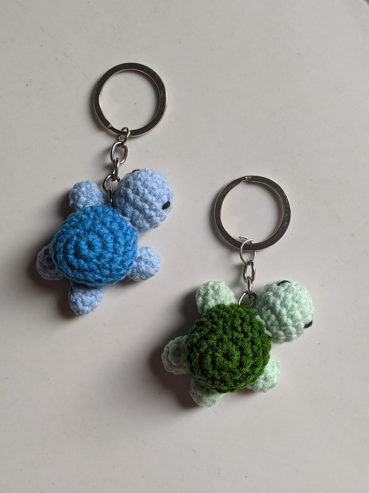 Clean side-by-side photo of two tiny crocheted turtle keychains, mint with dark green shell on the left and cream with denim blue shell on the right, both attached to silver keyrings on a white background