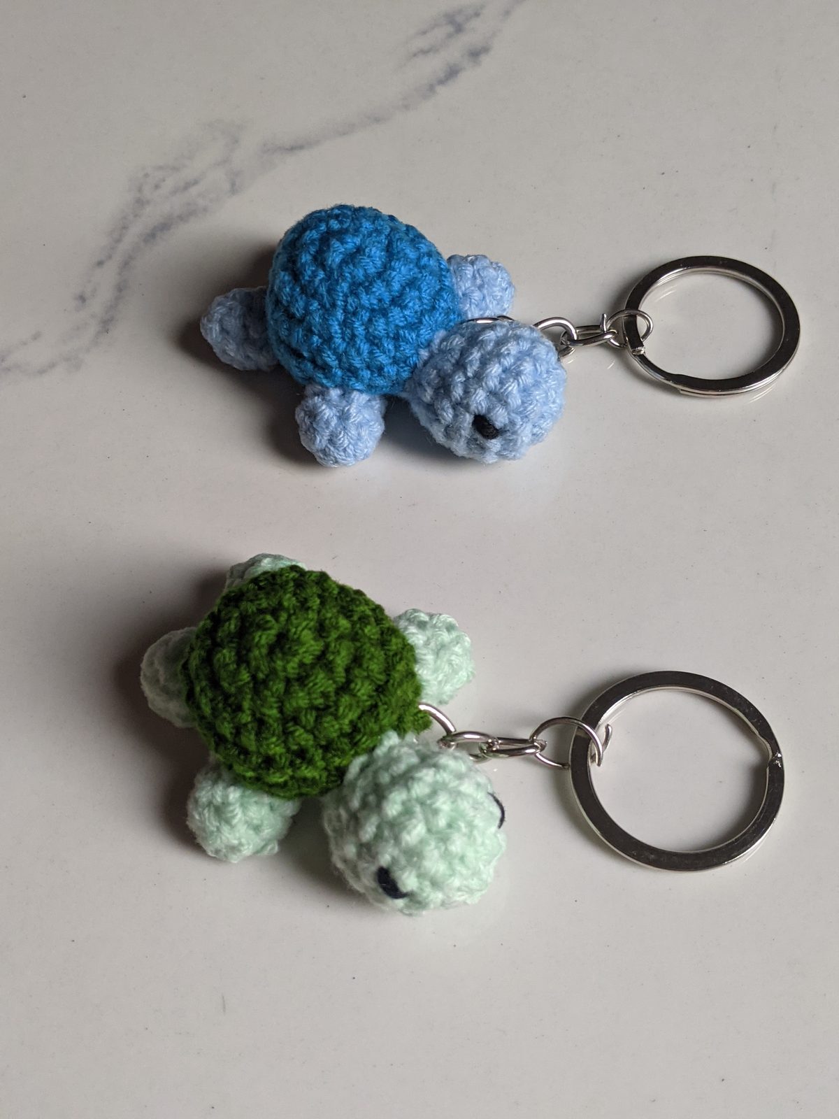 Two tiny crocheted turtle keychains, one mint and dark green and one blue, attached to silver keyrings, photographed from above