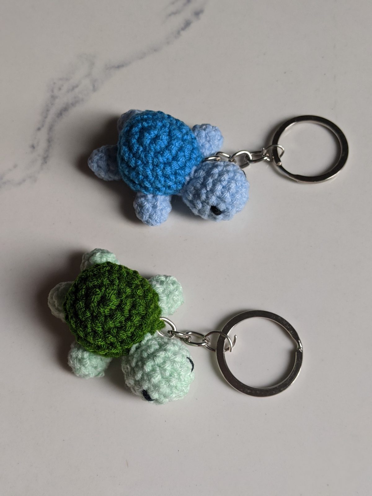 Two tiny crocheted turtle keychains, mint and dark green and blue, photographed side by side from a low angle showing both turtles clearly