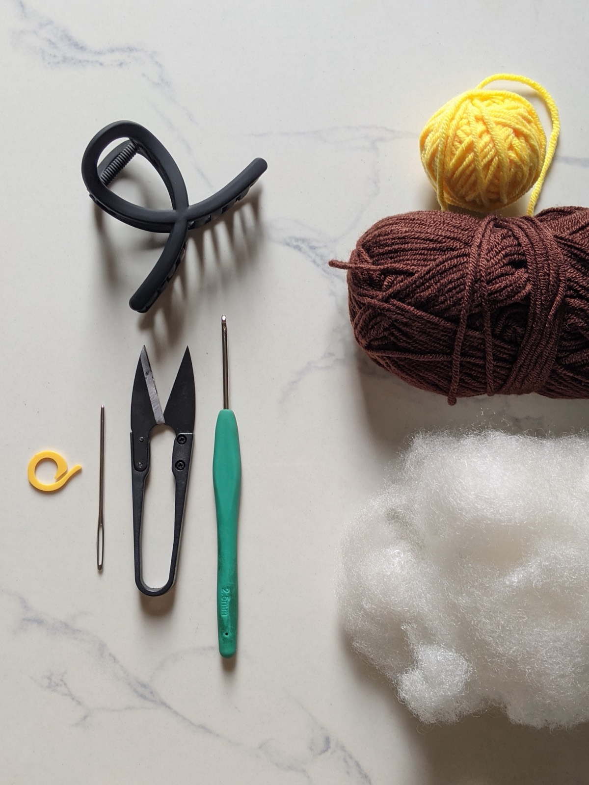 Flat lay of materials for the sunflower hair clip: yellow and dark brown milk cotton yarn, a black claw clip, a 2.5 mm crochet hook, scissors, a tapestry needle, and fiberfill stuffing