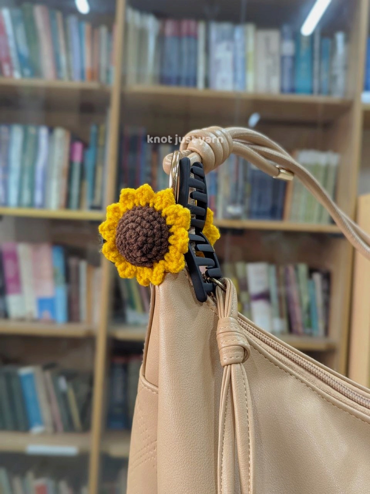 Sunflower clip attached to the strap of a tan shoulder bag in front of a bookshelf