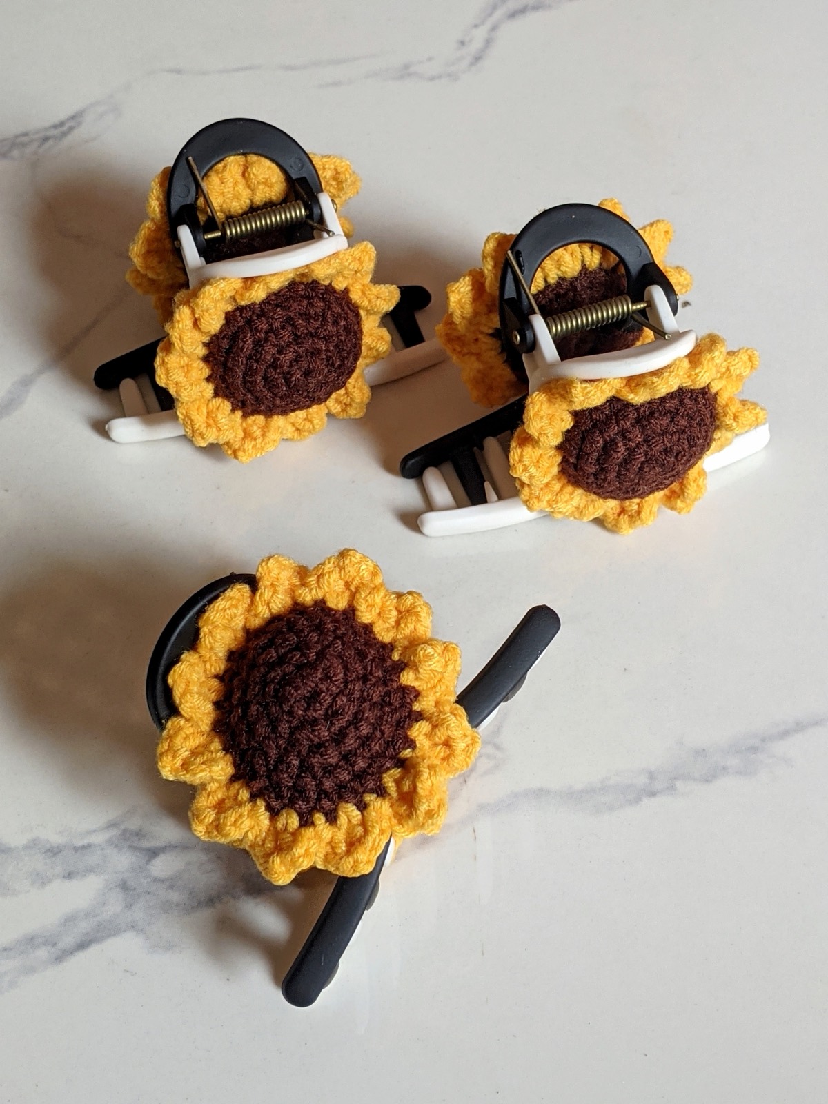 Three finished crochet sunflower claw clips arranged on a white marble surface, each with a brown center and yellow petals