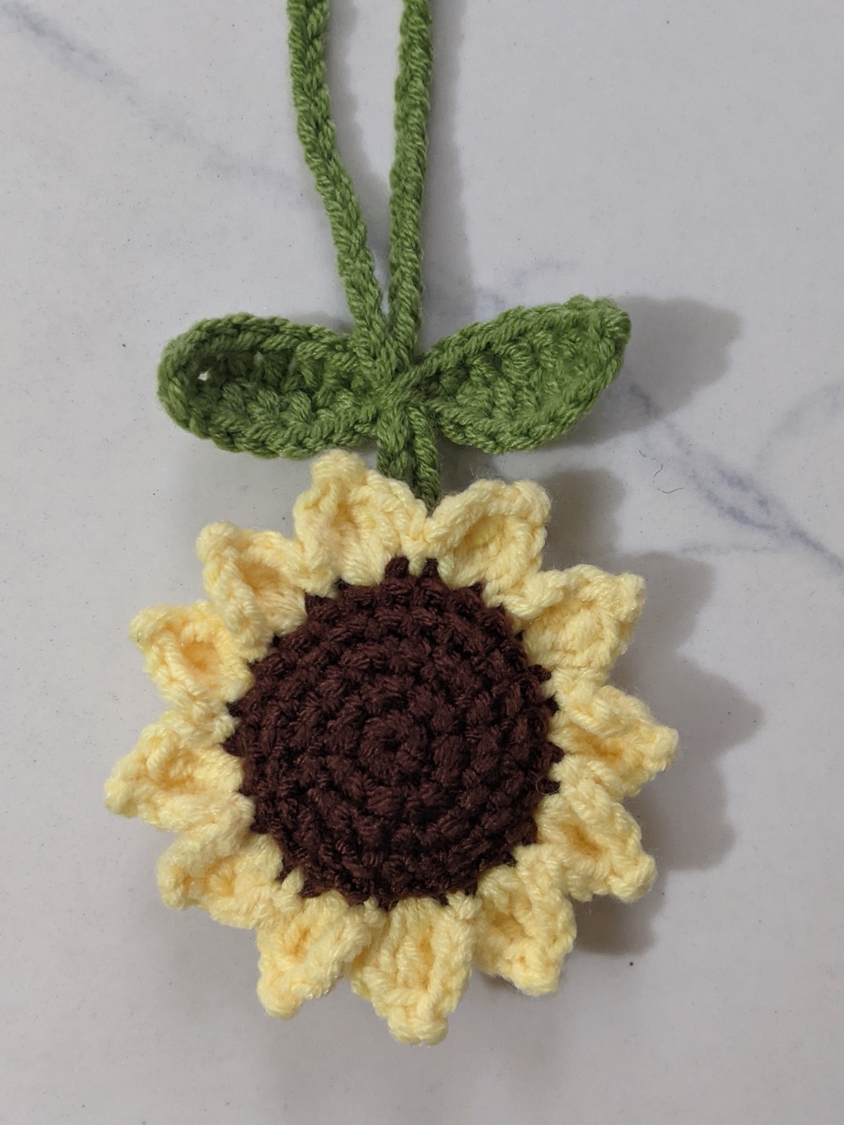 Finished crochet sunflower bag charm with green leaves and a hanging cord on a white marble surface