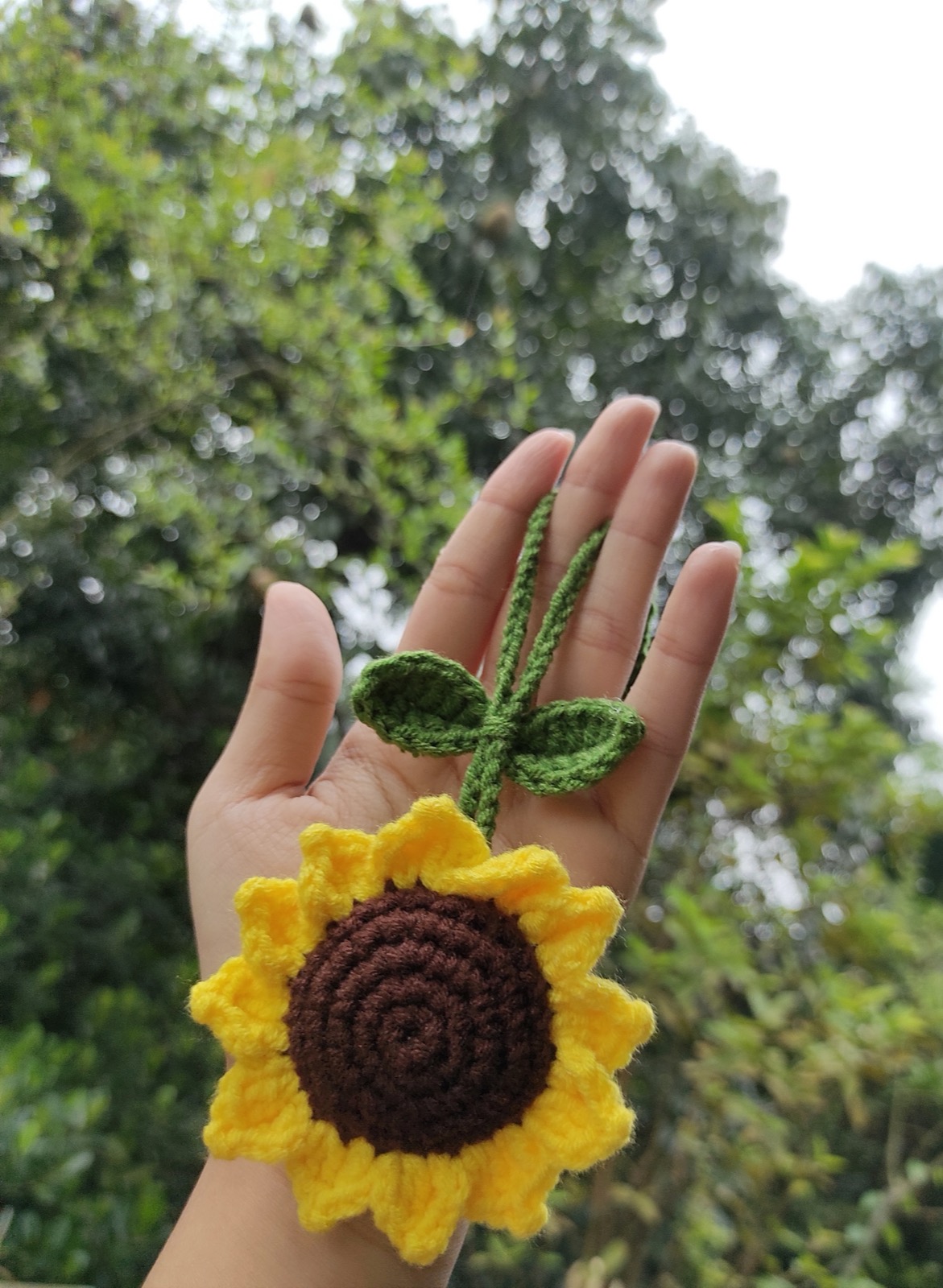 Hand holding the finished crochet sunflower charm with leaves outdoors, showing its real size
