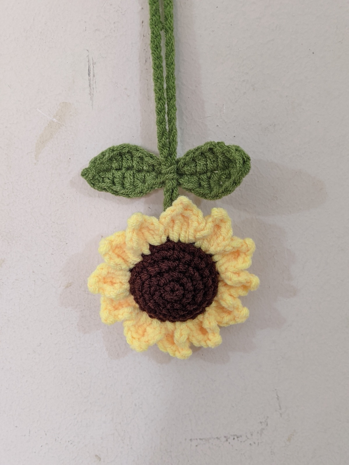 Crochet sunflower charm with leaves and a green hanging cord displayed against a pale wall