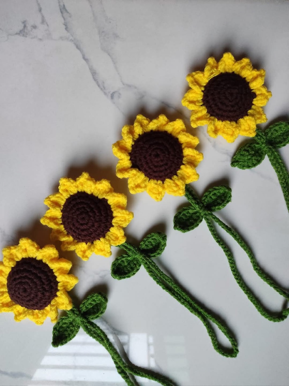 Four golden crochet sunflowers with green leaves and stems arranged on white marble