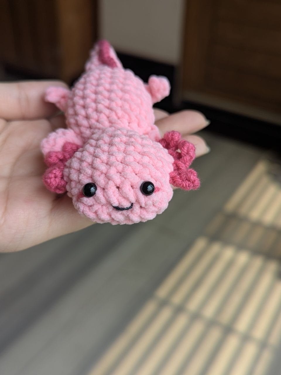 Front view of a finished pink crochet axolotl with dark pink frilly gills and an embroidered smile, held in a hand