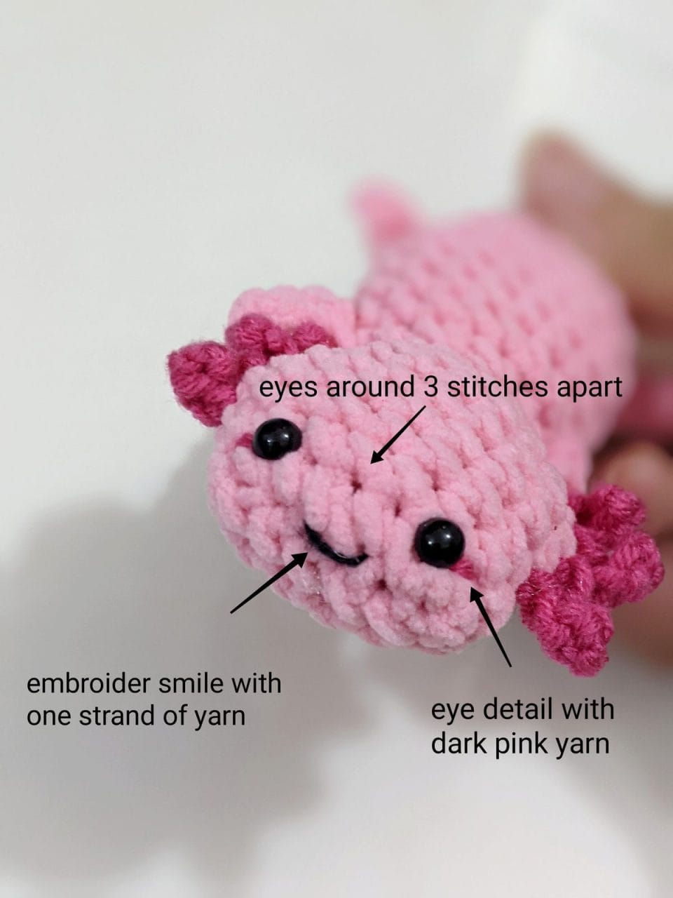 Finished crochet axolotl with safety eyes placed, dark pink eye accents and a black embroidered smile