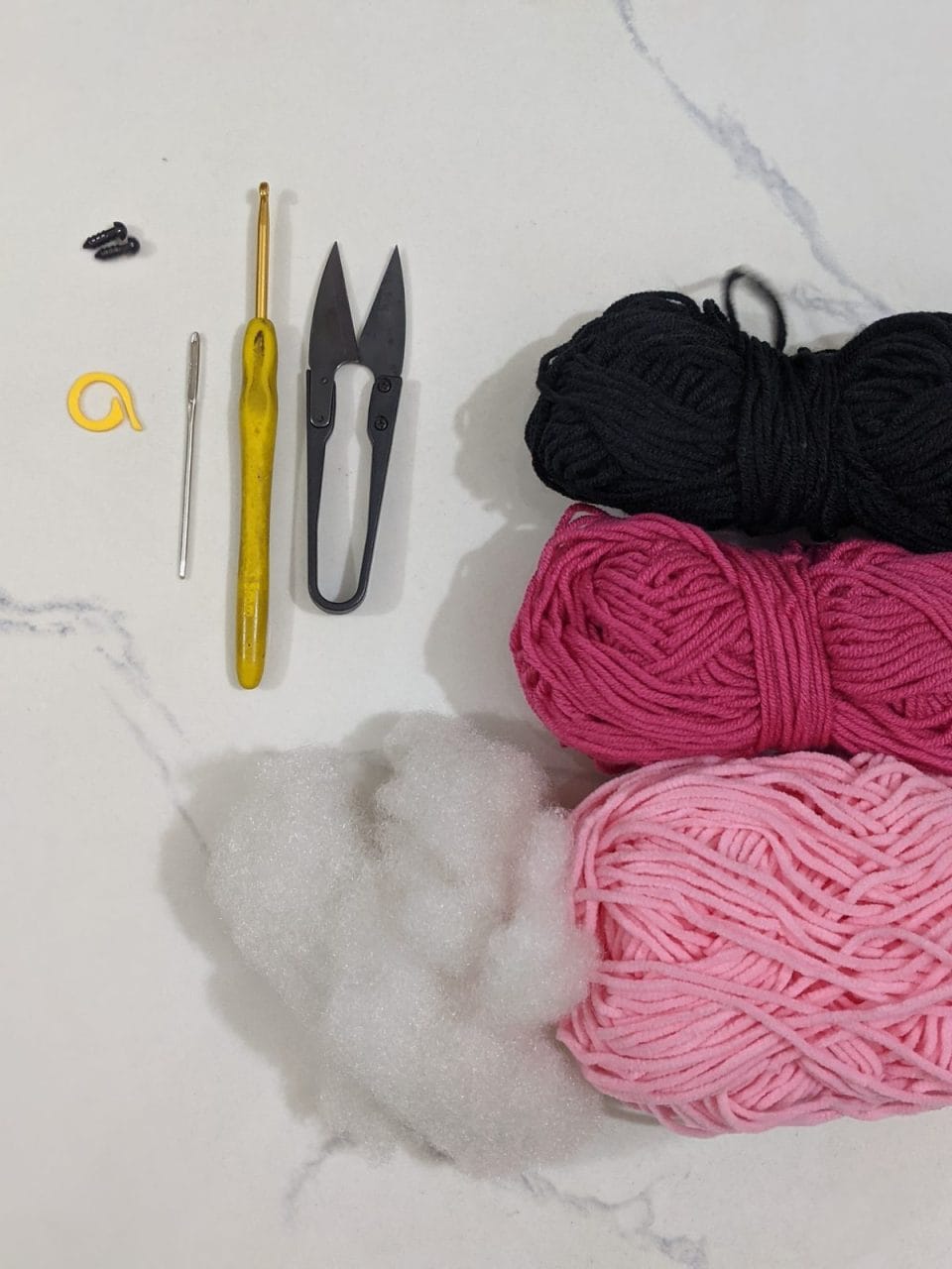 Flat lay of axolotl crochet materials: light pink plush yarn, dark pink and black milk cotton yarn, 3.5 mm hook, safety eyes, fiberfill, scissors, yarn needle and stitch marker