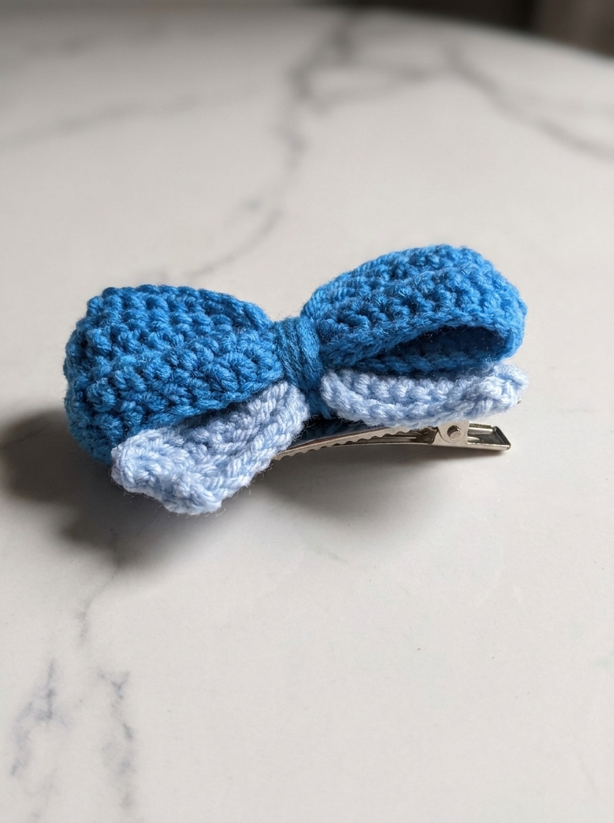 Crocheted blue and light-blue puffy bow attached to a metal single-prong alligator hair clip, resting on a white marble surface