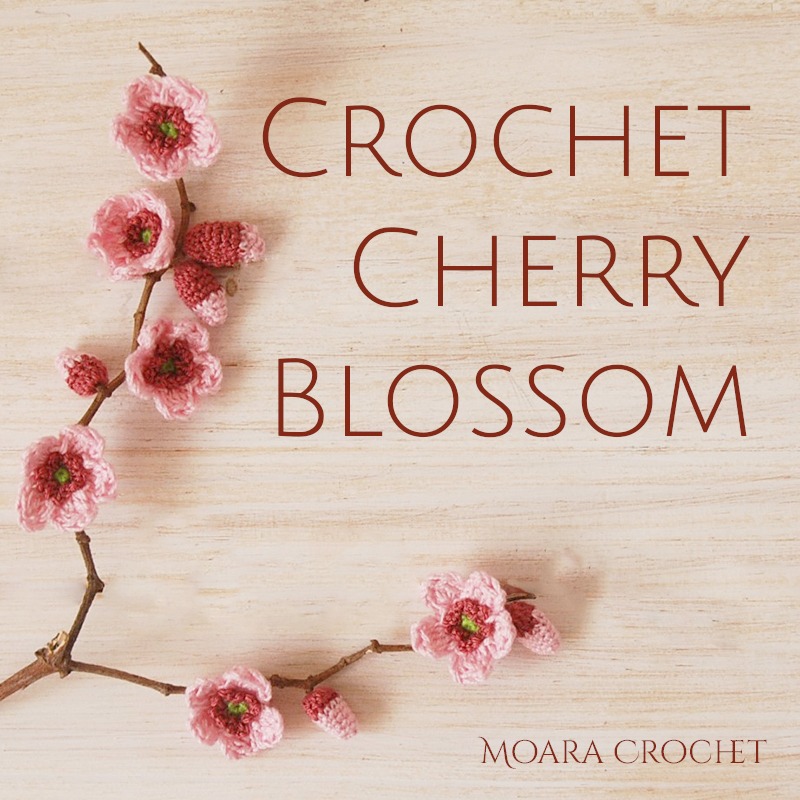 Small pink crochet cherry blossom flowers attached along a real twig branch on a pale wood background, from Moara Crochet