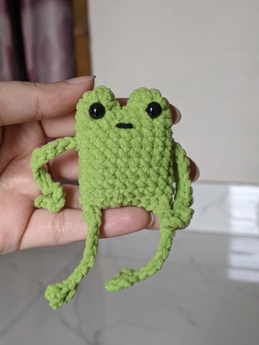 A hand holding the finished green crochet leggy frog showing its small palm-sized scale