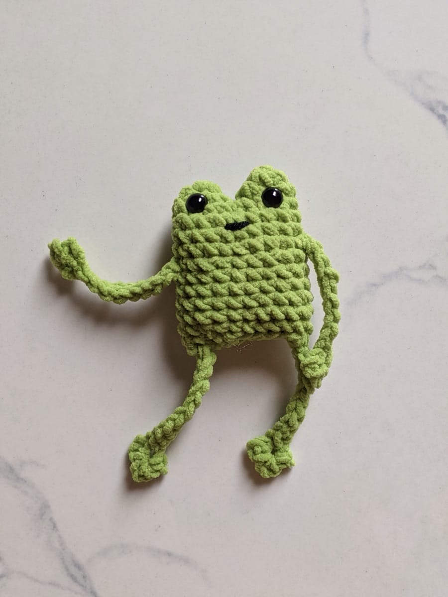Finished green crochet leggy frog amigurumi with long floppy chain arms and legs and black safety eyes on a marble surface