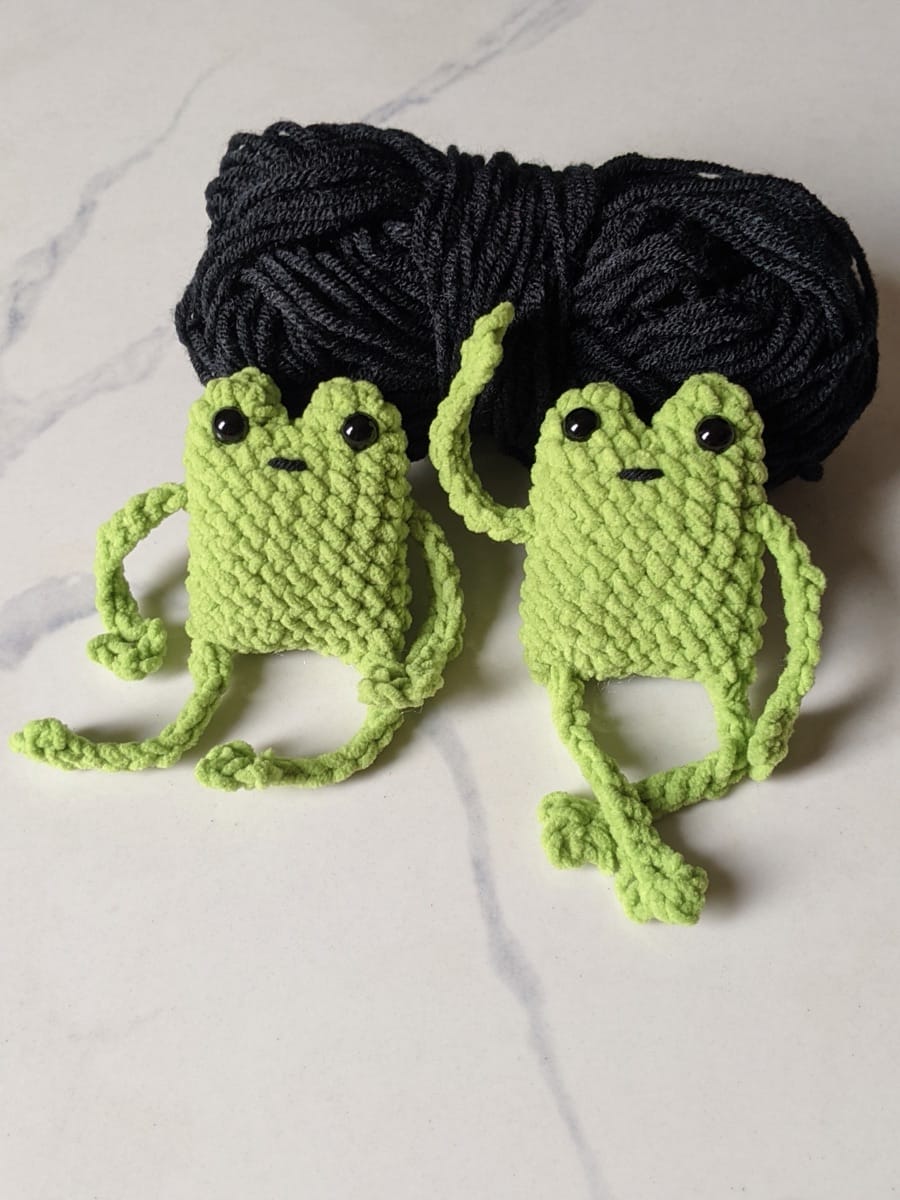 Two green crochet leggy frogs sitting side by side with a black skein of yarn behind them