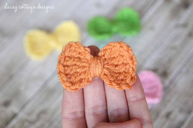 A small orange crochet bow with a brown bead center balanced on two fingers, with yellow, green and pink bows blurred in the background