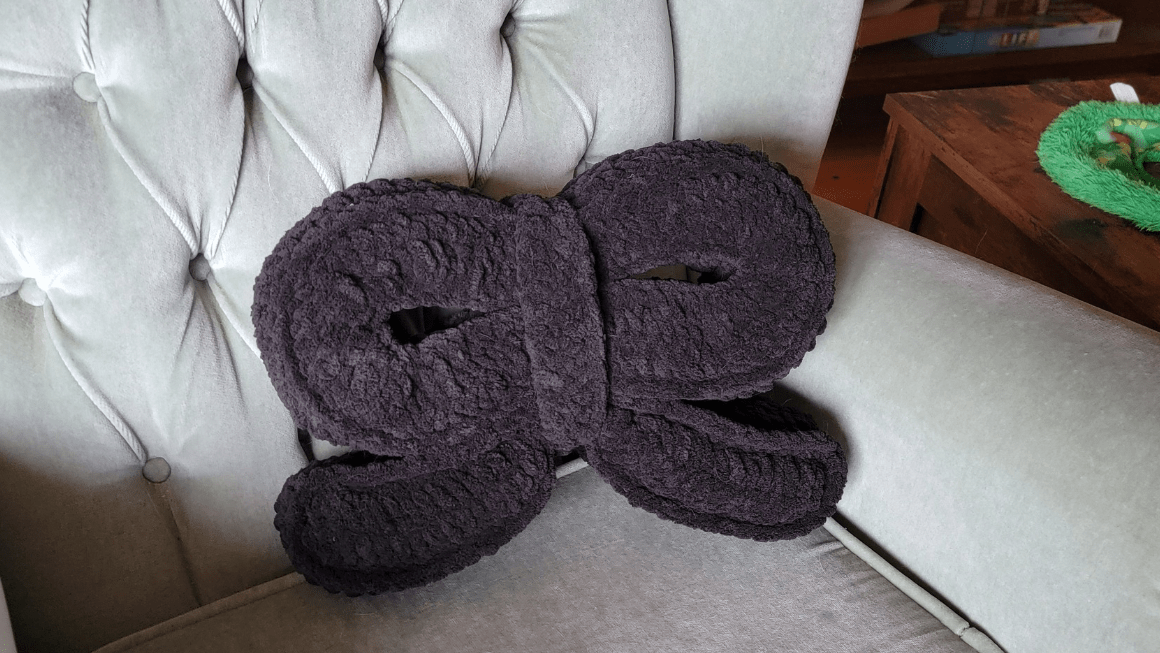 A large charcoal crochet bow pillow made in chunky blanket yarn, resting on a pale grey tufted armchair
