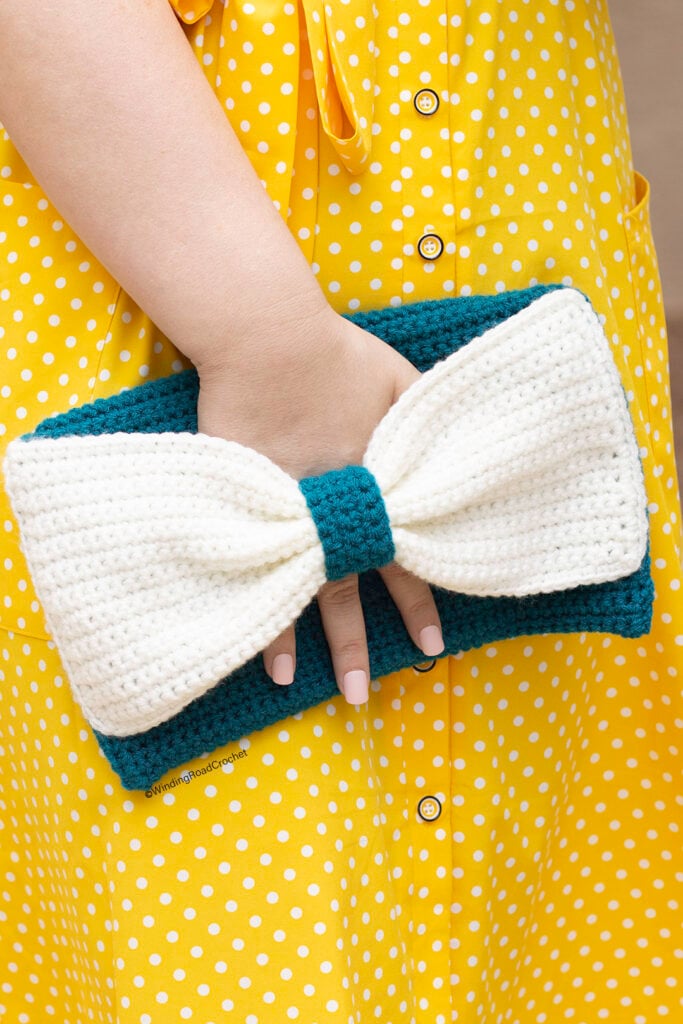 A teal crochet clutch with a large cream bow across the front, held in one hand against a yellow polka dot dress