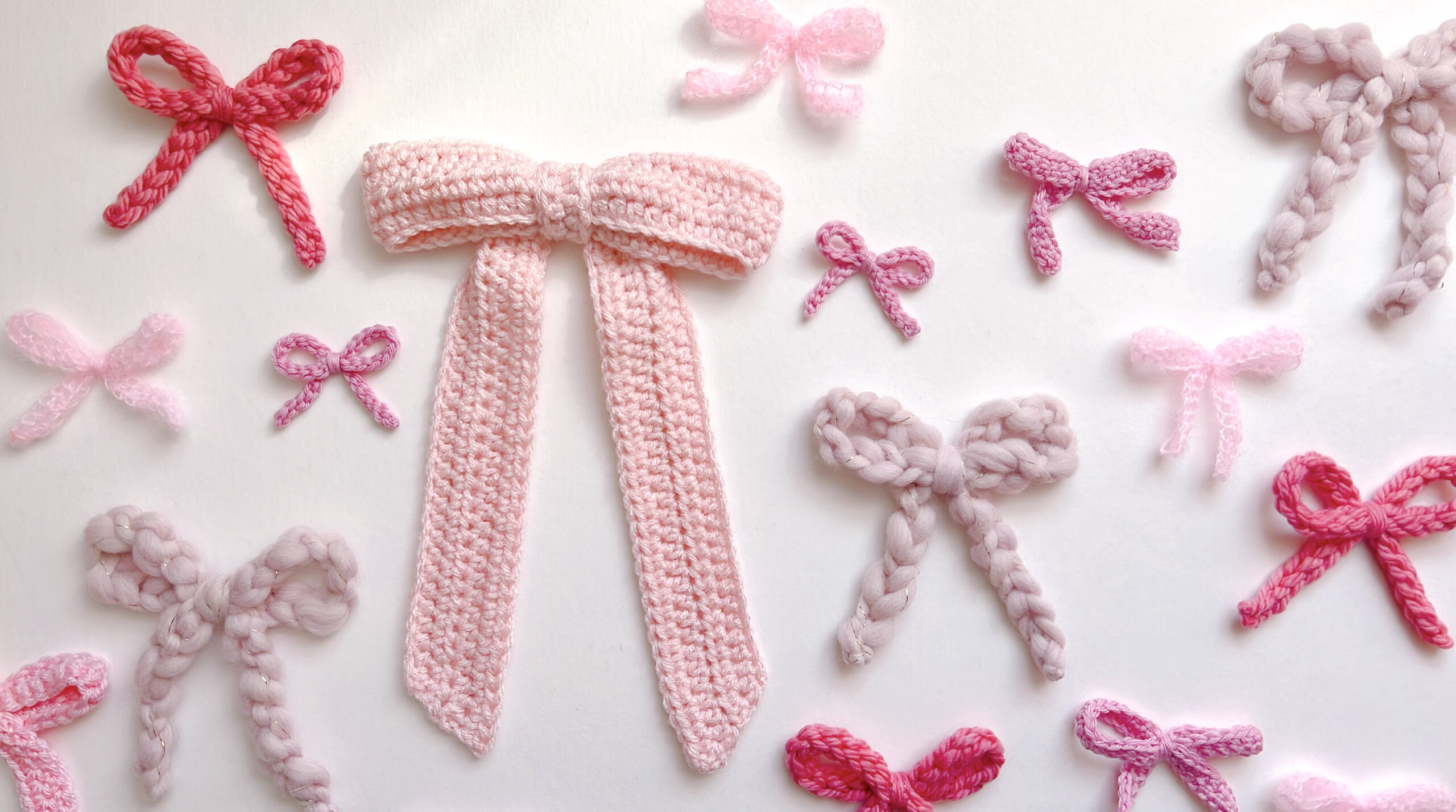 Many small crochet bows in pink, rose and blush laid out on white, worked in bulky wool, worsted acrylic and thin mohair, with one large pink bow with long tails in the center