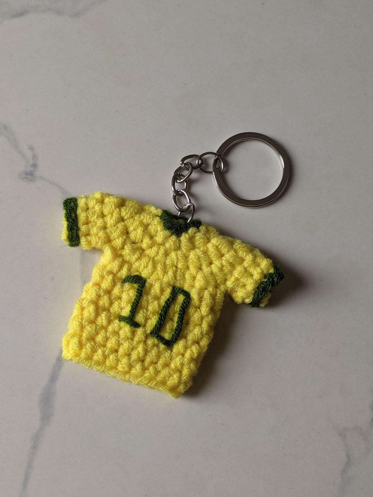 The finished yellow crochet Brazil jersey keychain with green neck and cuff trim, hanging from a silver chain and split ring on a marble counter
