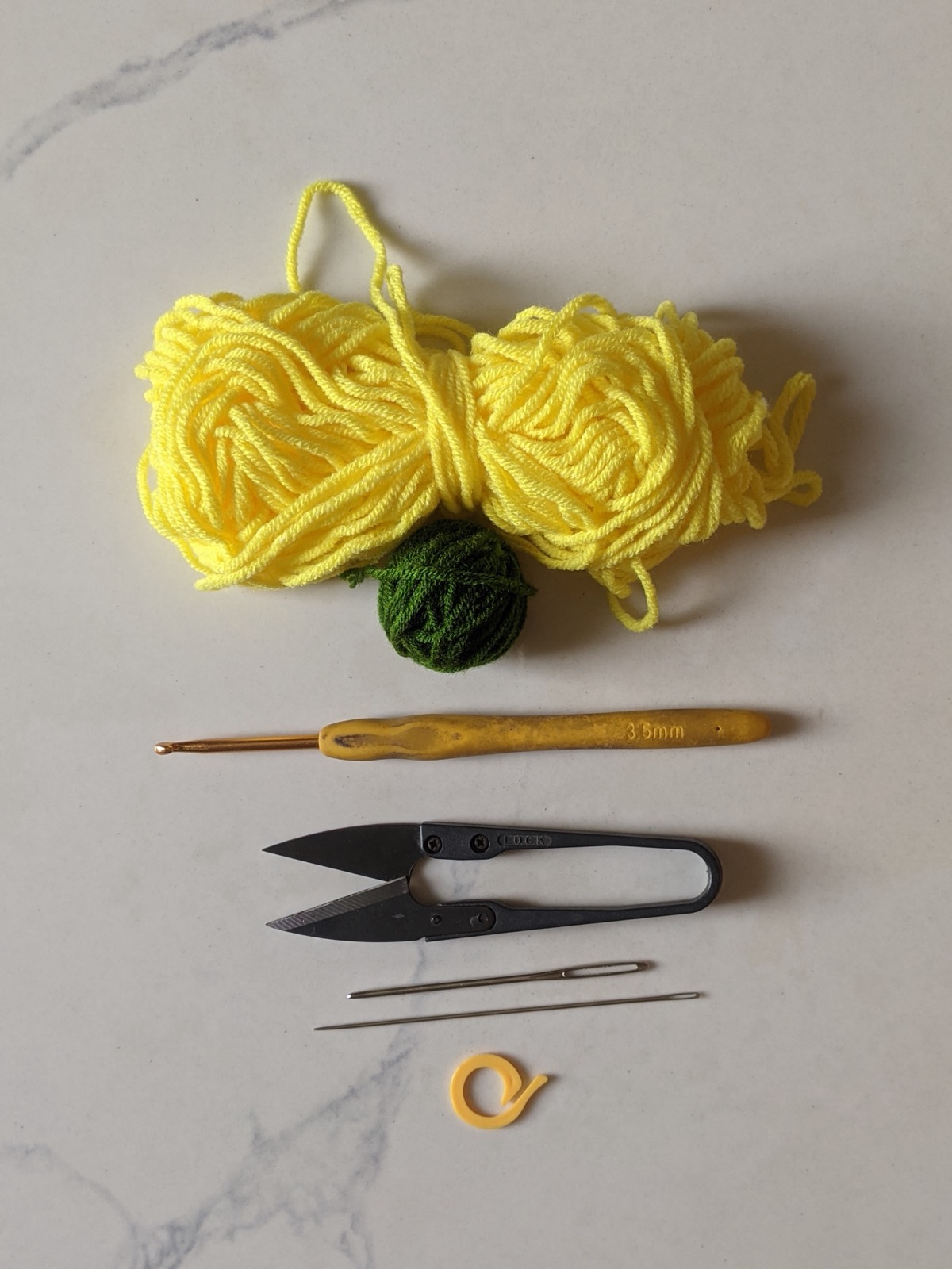 Flat lay of a skein of yellow milk cotton yarn, a small ball of green, a 3.5 mm crochet hook, thread snips, two tapestry needles and a stitch marker