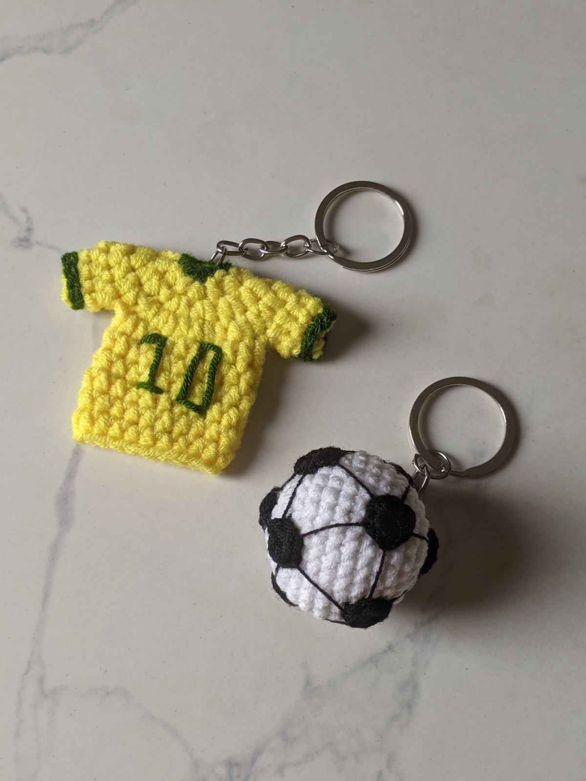 The yellow crochet jersey keychain and a white and black crochet soccer ball keychain, both on silver chains and split rings, laid on a marble counter