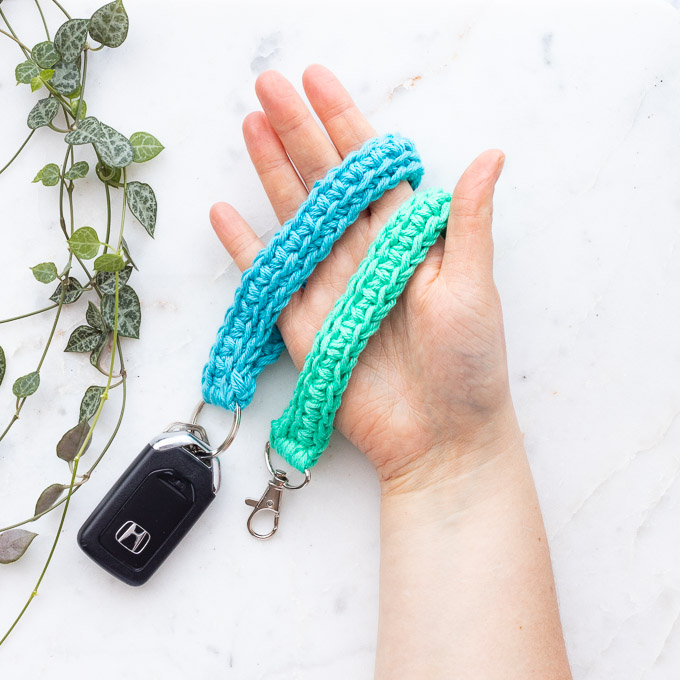 Two crocheted wristlet key straps in teal and mint held in an open hand on white marble, one attached to a car key