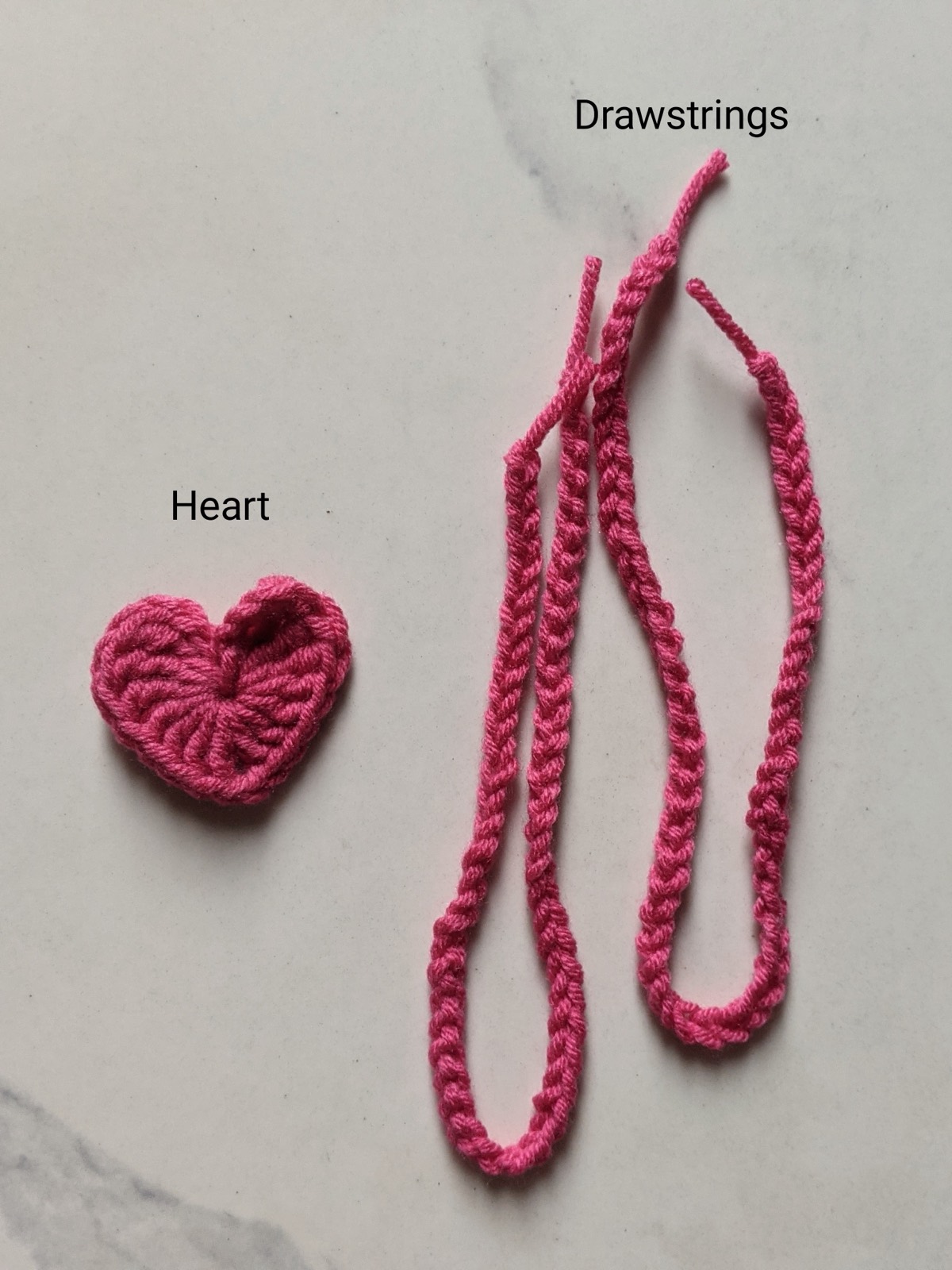 The two dark pink crochet drawstrings laid out beside the small crochet heart motif before assembly