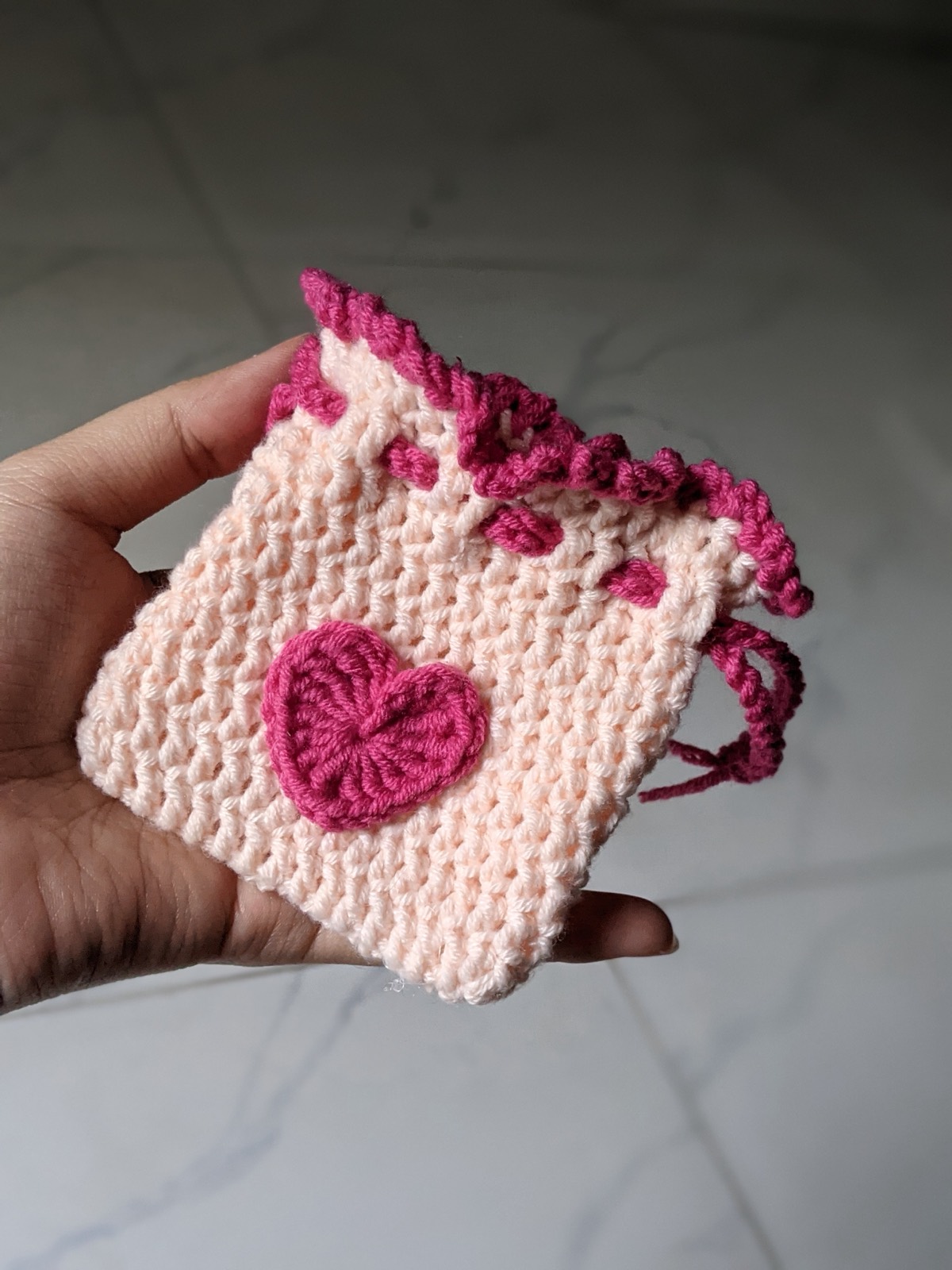 A pink crochet mini pouch held in a hand to show its size, cinched closed with two dark pink drawstrings