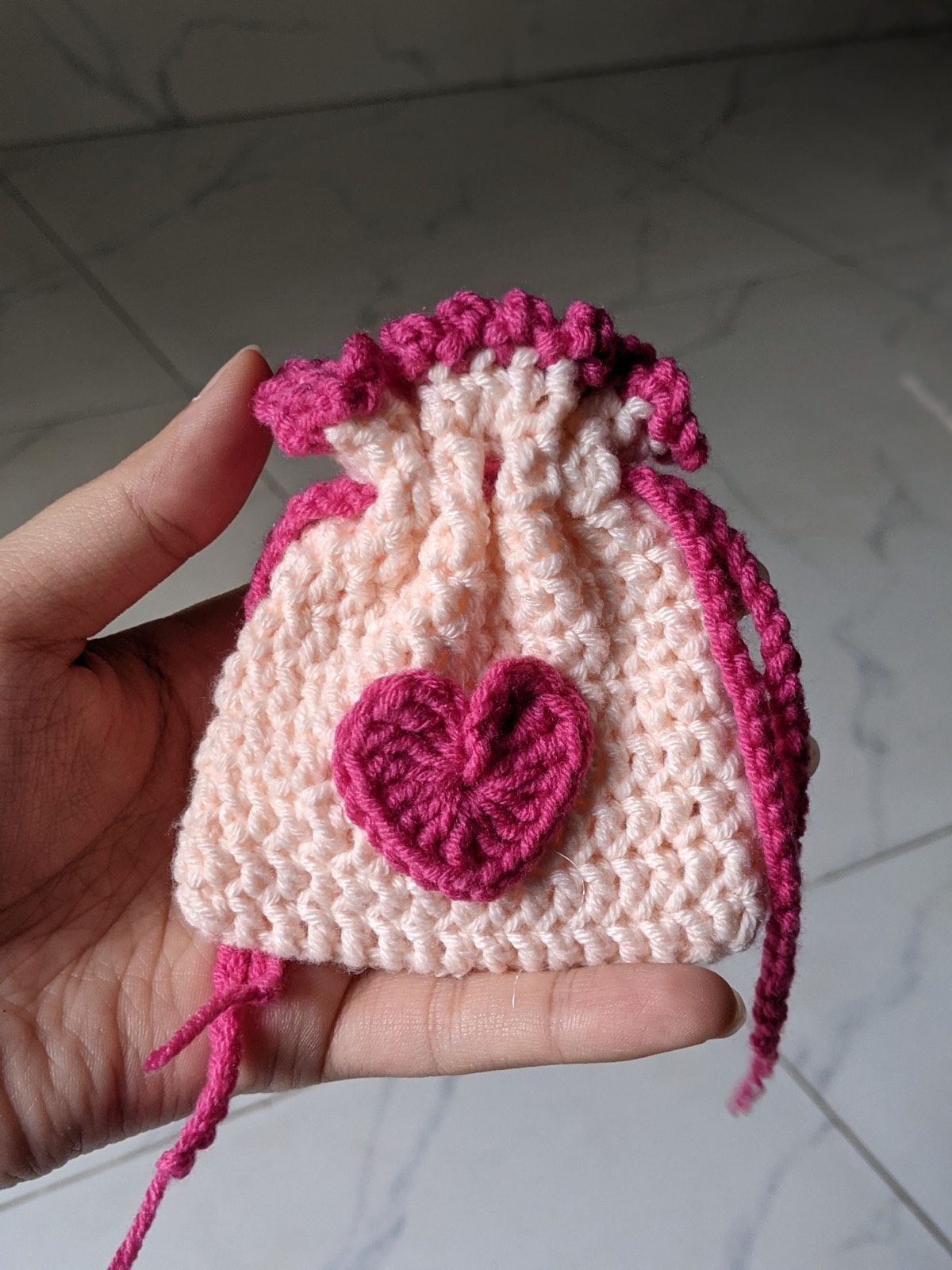 A hand holding a small pink crochet drawstring pouch closed shut, showing it fits easily in a palm