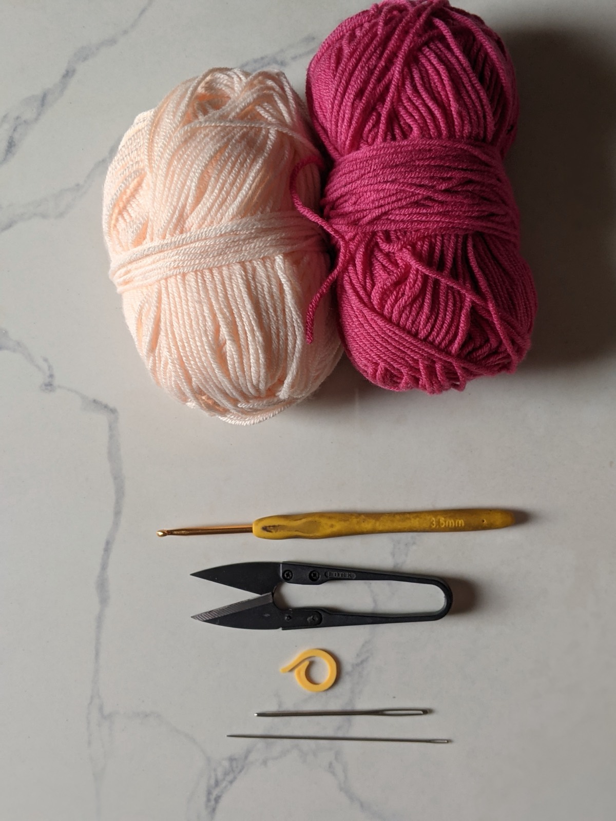 Flat lay of materials for the crochet mini pouch: light pink and dark pink cotton yarn balls, a 3.5 mm hook, snips, a stitch marker and two yarn needles