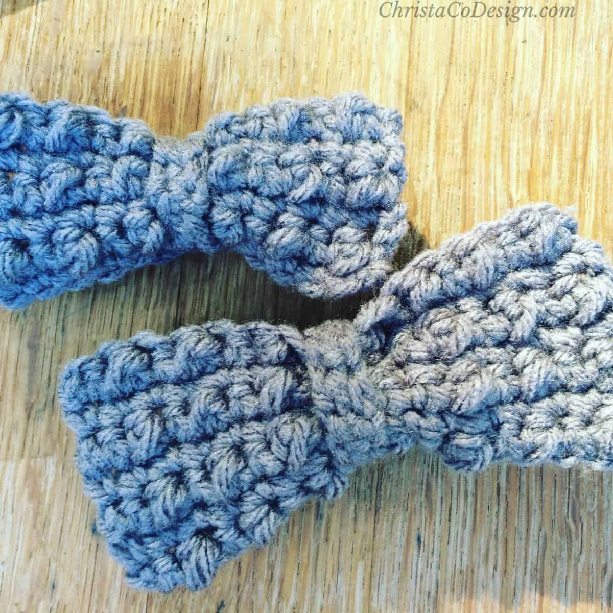 Two blue textured crochet bow ties in child and baby sizes lying on a light wooden surface
