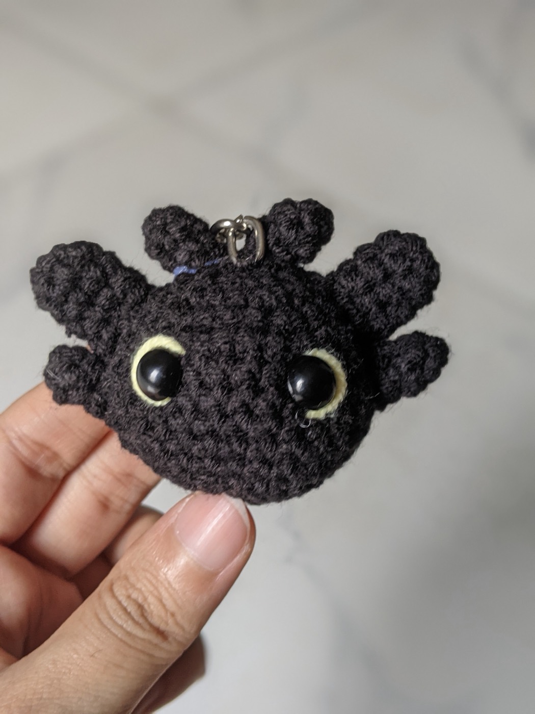 Close up of the black crochet dragon keychain held between finger and thumb, showing six head fins and yellow embroidered rings around the safety eyes