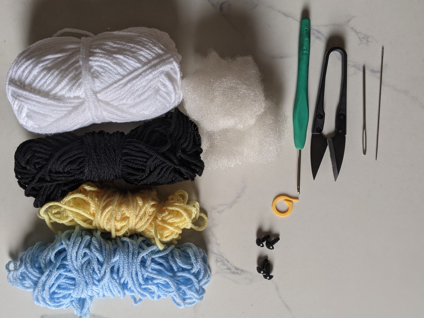 Flat lay of crochet supplies including white, black, yellow and light blue yarn, a 2.5 mm hook, fiberfill, snips, two yarn needles, a stitch marker and four black safety eyes