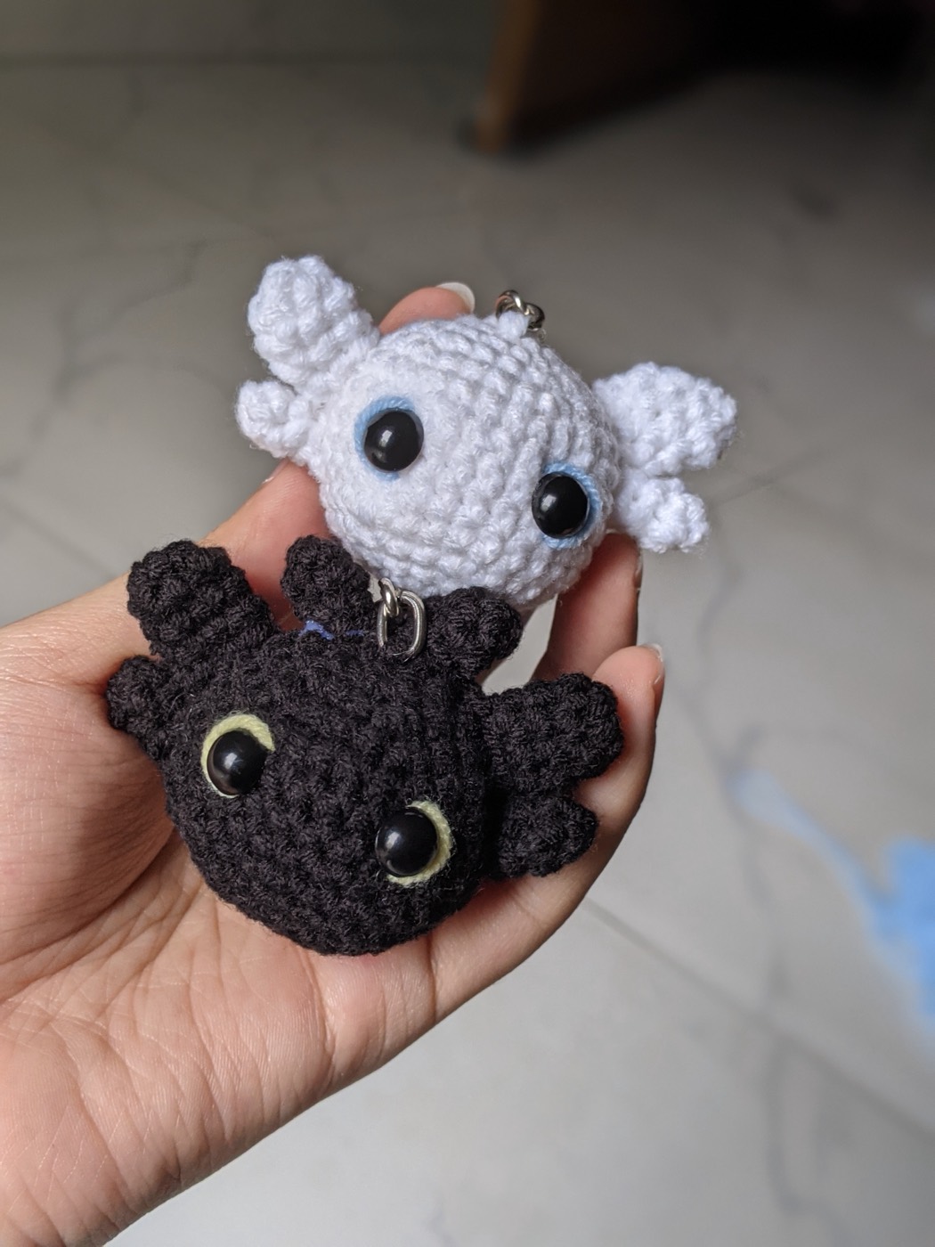 A white crochet dragon head and a black crochet dragon head held together in a palm, both on small keyring chains
