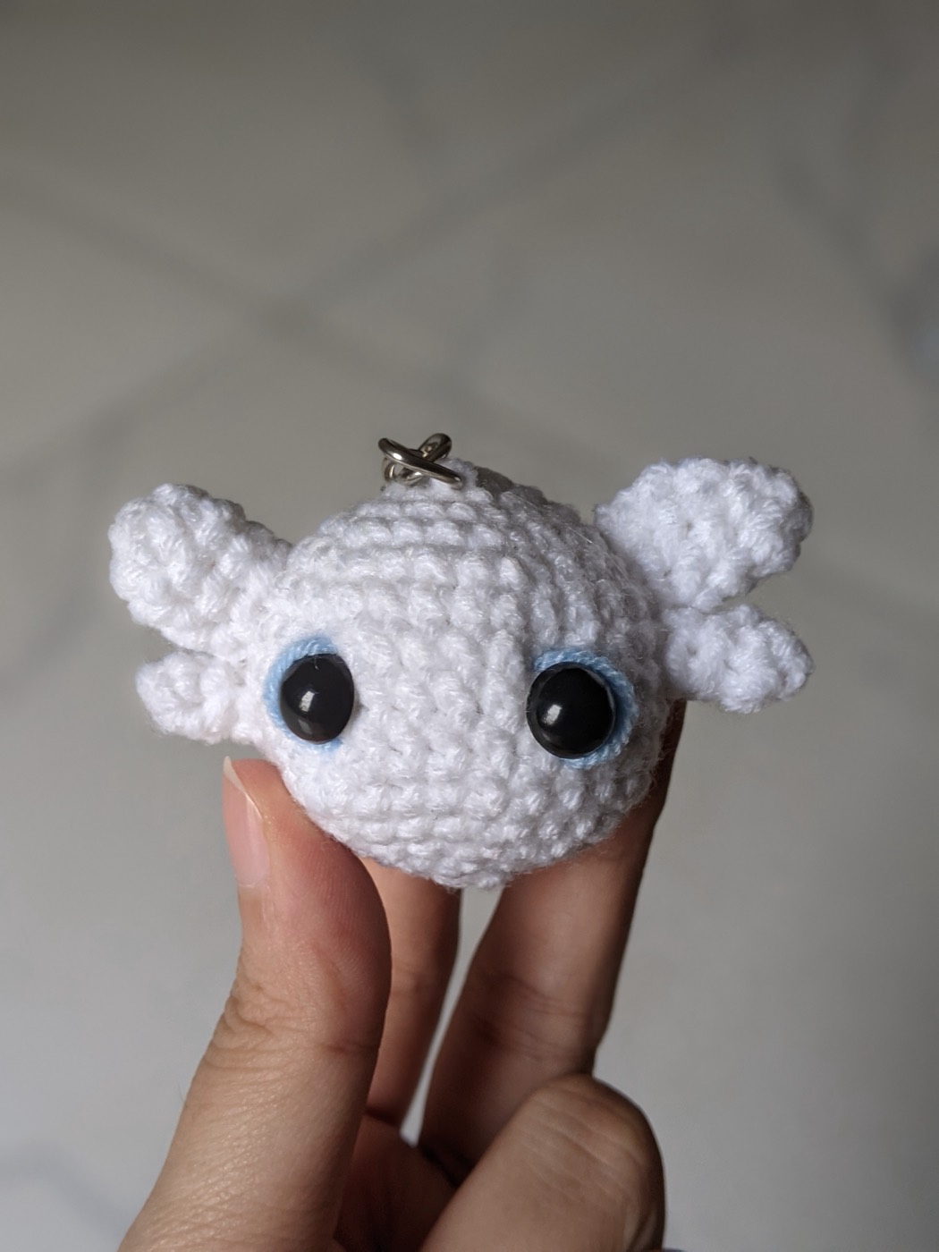 Close up of the white crochet dragon keychain resting on a fingertip, showing four white head fins and light blue embroidered rings around the safety eyes