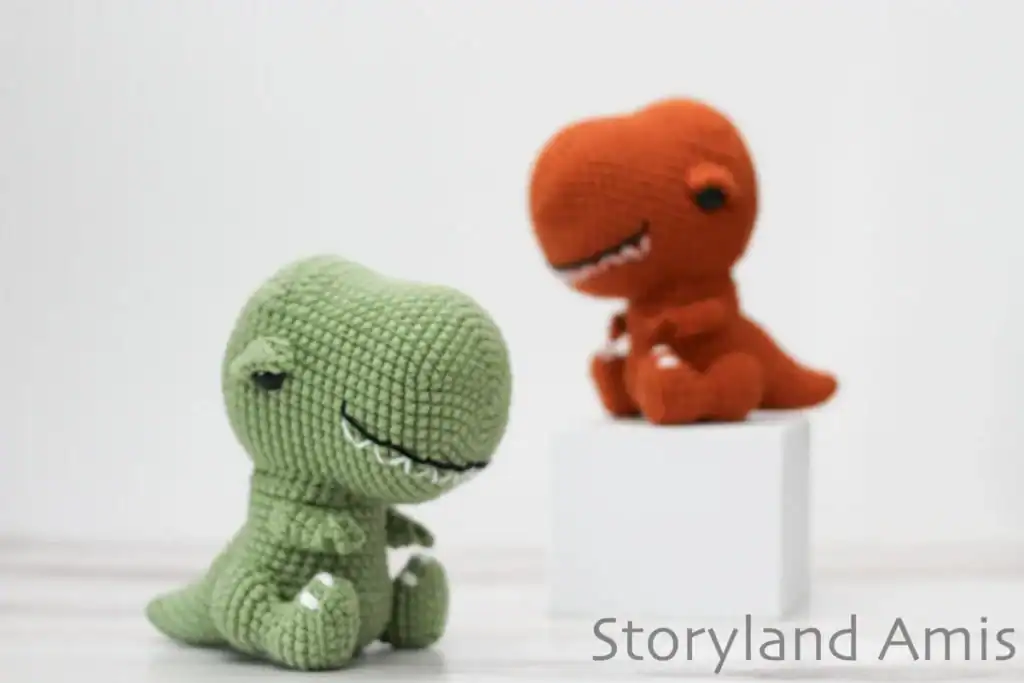 Two crochet T-rex amigurumi, one sage green and one orange, sitting with embroidered teeth and tiny arms, from Storyland Amis