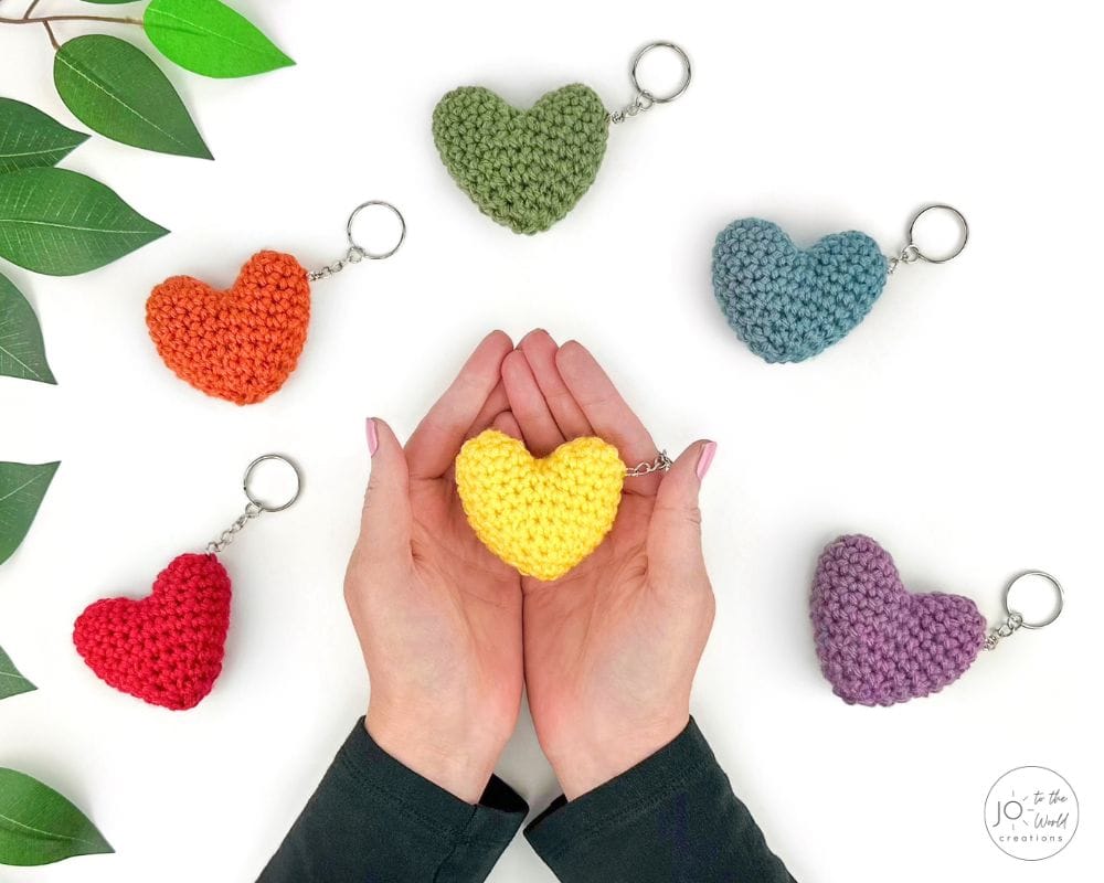 Seven small crocheted heart keychains in different colors on silver key rings, with a yellow one held in two hands