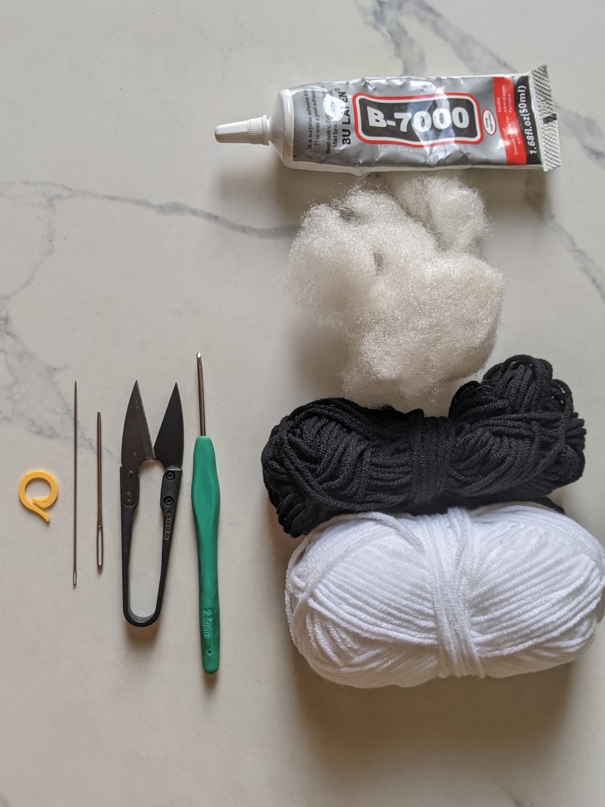 Flat lay of crochet supplies including white and black yarn, a 2.5 mm hook, yarn needles, snips, a stitch marker, fiberfill and a tube of craft adhesive